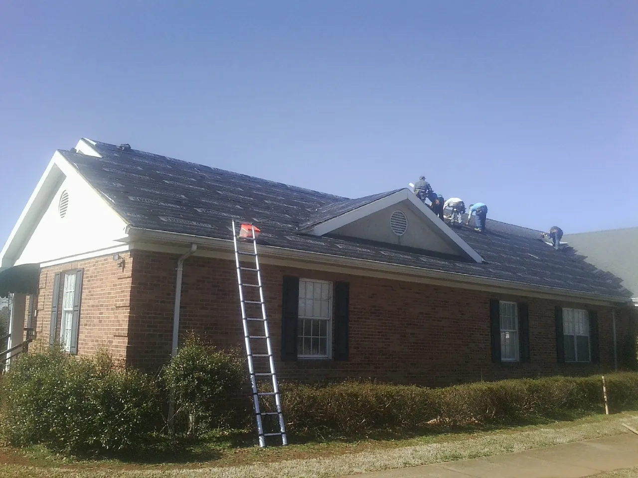 Crew tearing shingles off a brick ranch roof with a ladder set — Emergency Roof Repair work by our crew in Ranchos De Taos