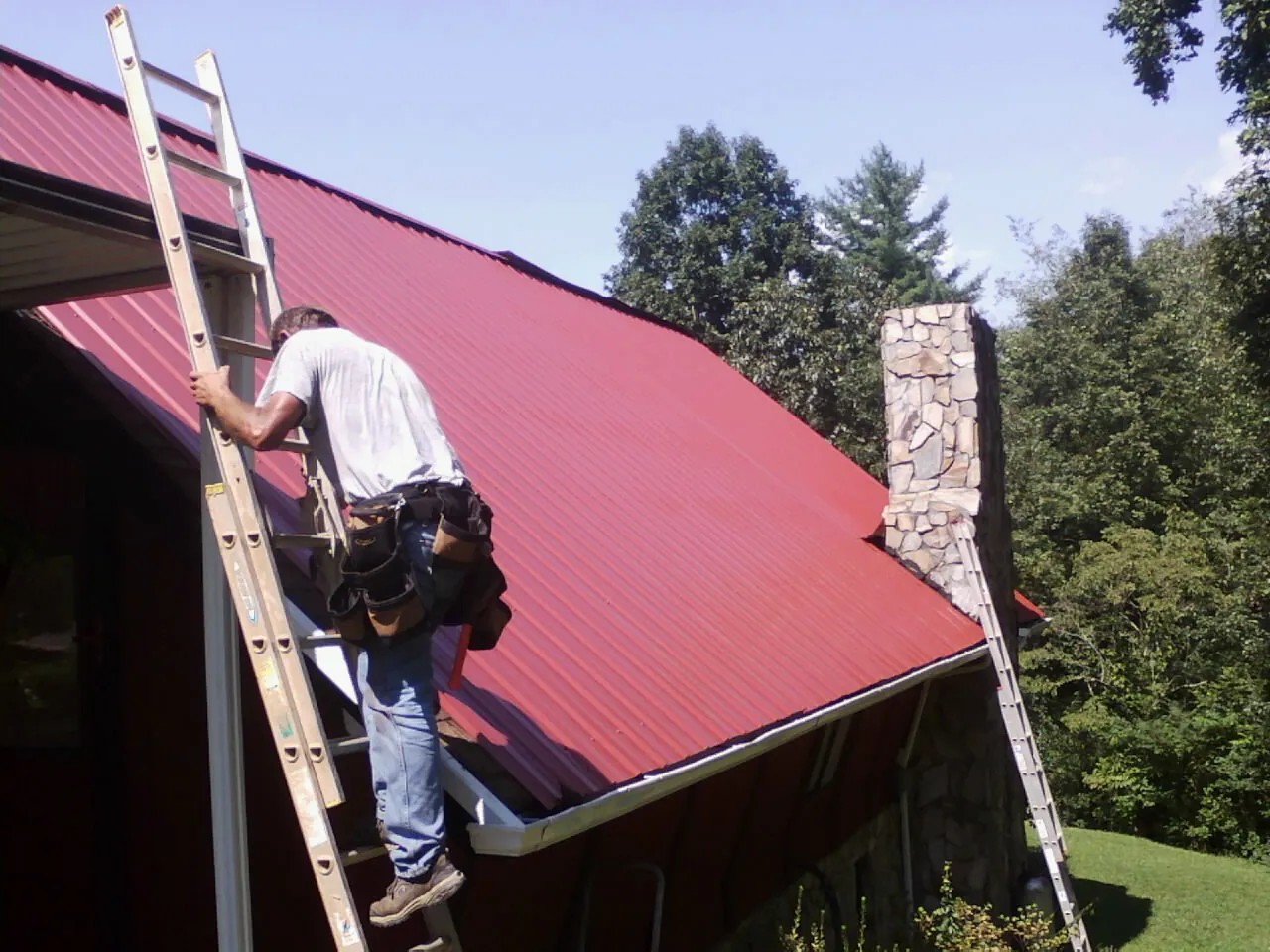 Roofer on a ladder beside a red metal roof and stone chimney — Soffit & Fascia Repair by Roofing Brothers in Ranchos De Taos